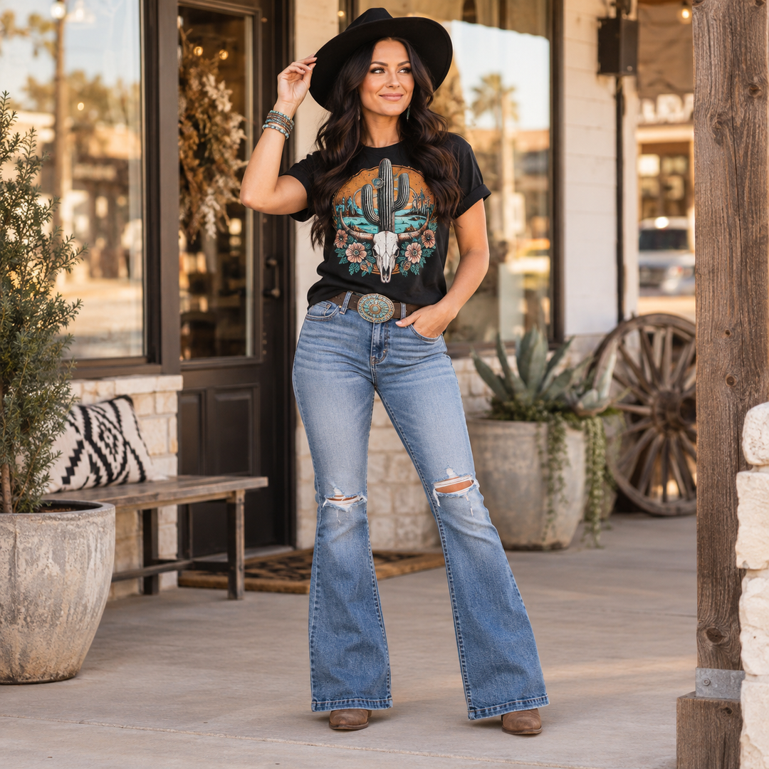 Dark-haired woman in a bold Western graphic tee, flared jeans and cowboy hat outside a boutique