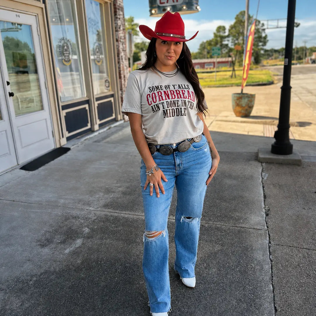 Full-length outdoor view of the heather tan cornbread saying graphic tee styled with jeans and a red cowboy hat