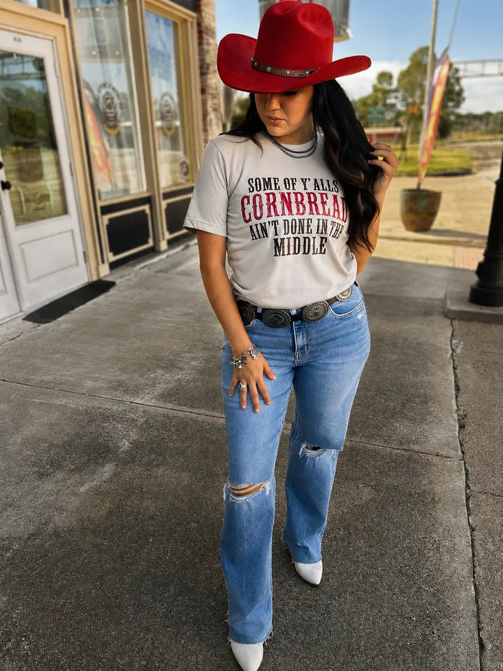 Outdoor three-quarter view of the heather tan cornbread saying graphic tee styled with jeans and Western accessories