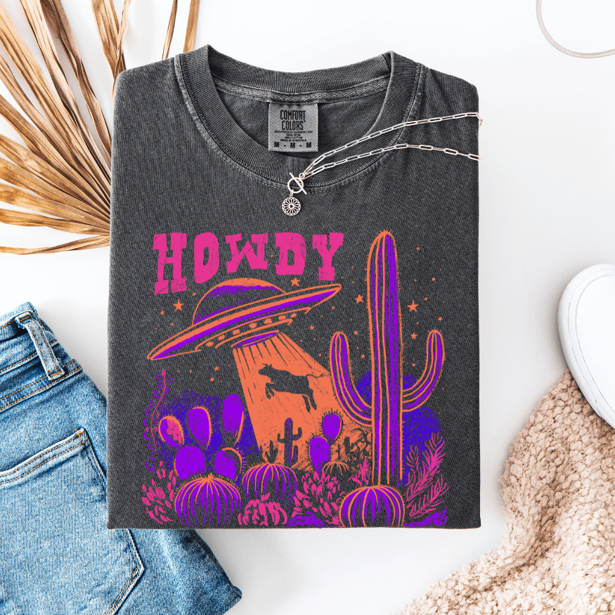 Women wearing charcoal gray unisex tee with neon pink "Howdy" text, UFO abducting cow, and purple cactus graphics
