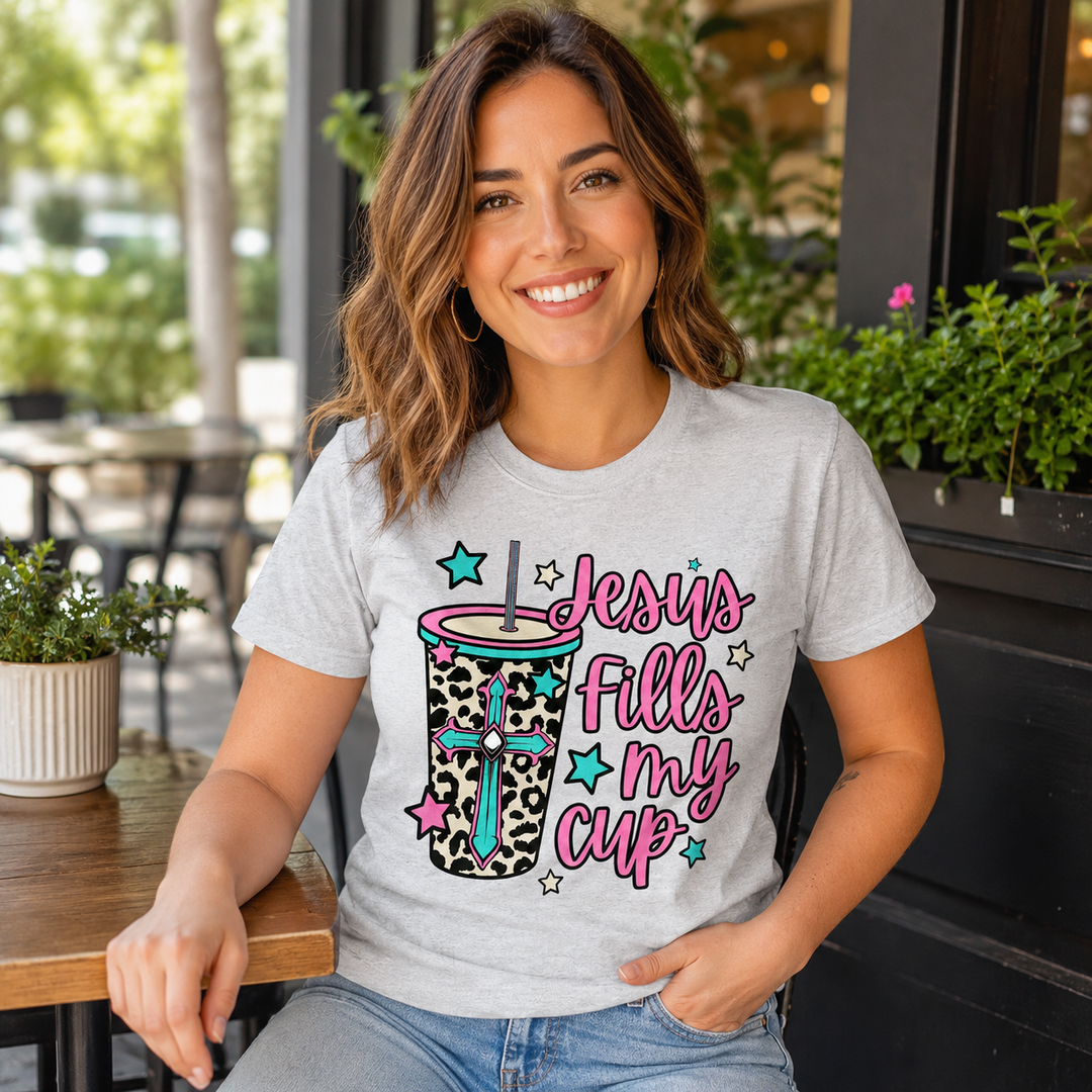 Woman wearing the Jesus Fills My Cup leopard tumbler faith graphic tee at a coffee-shop patio