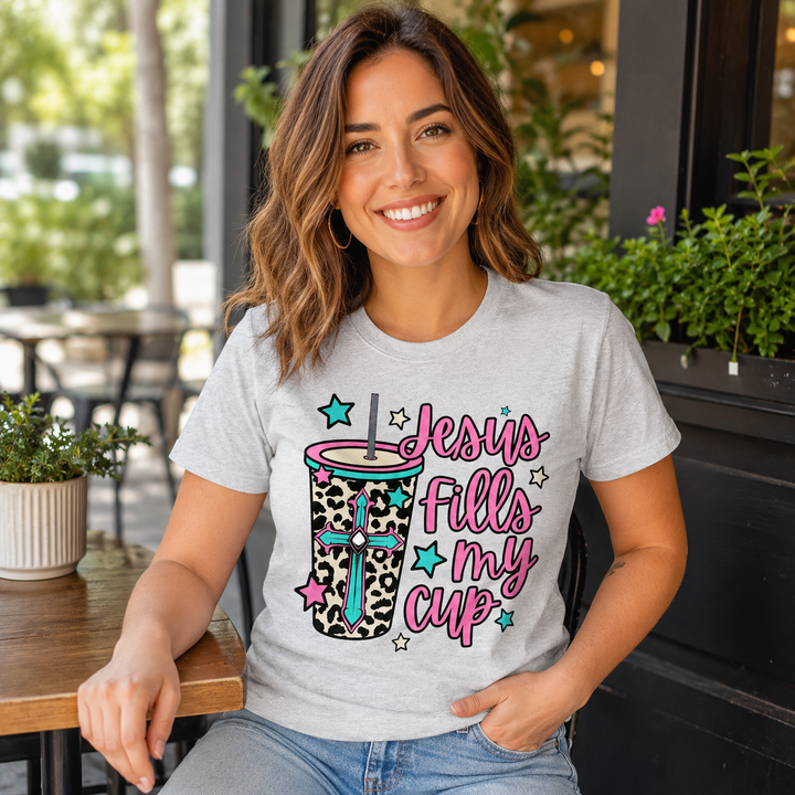 Woman wearing the Jesus Fills My Cup leopard tumbler faith graphic tee at a coffee-shop patio