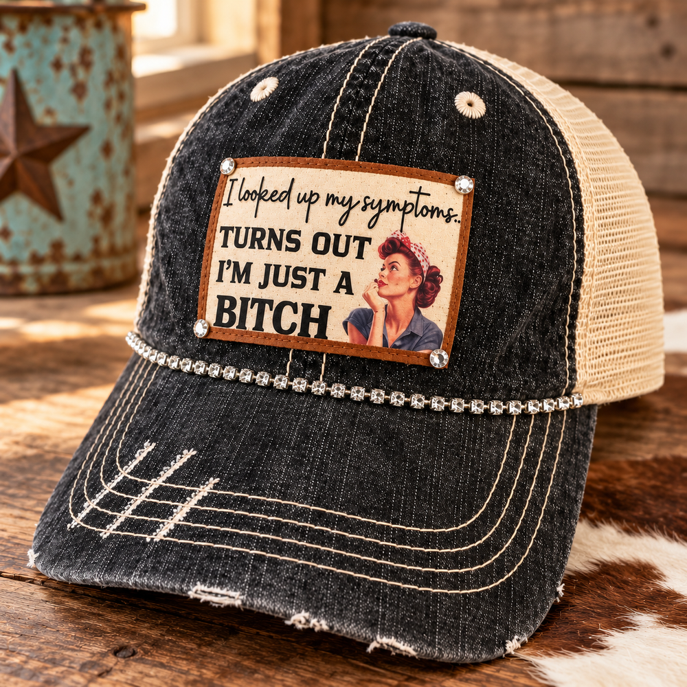 Close three-quarter view of Just a B*tch Trucker Hat with distressed black front, tan mesh, vintage patch, and rhinestone trim